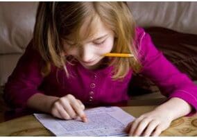 A young girl doing homework with a pencil in her mouth.